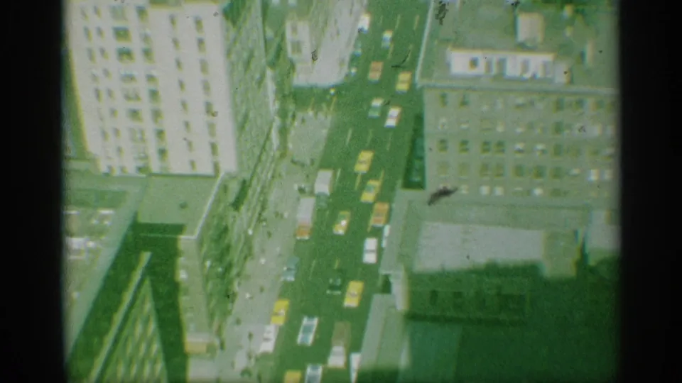 1959 New York aerial-style home-movie frame showing a vertical street between tall buildings, visible cars, rooftops, and green-yellow film color.