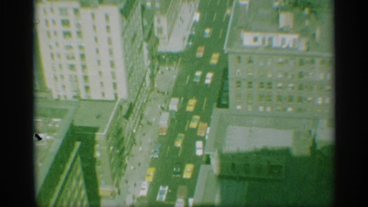 1959 New York overhead street view with traffic, yellow taxis, sidewalks, and high-rise buildings seen through 8mm film grain.