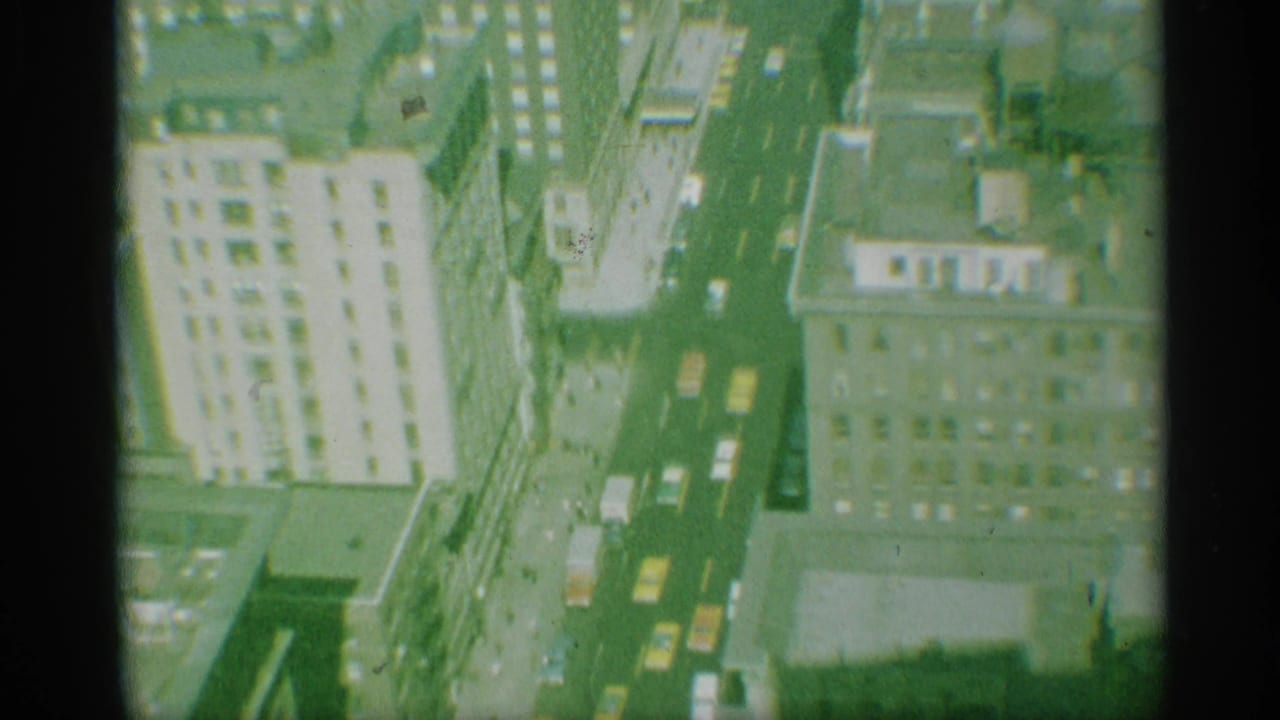 Overhead 1959 New York street scene showing cars and yellow cabs moving between tall commercial buildings.