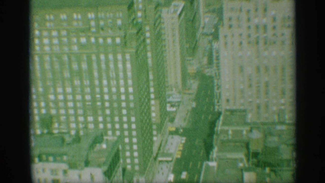 1959 New York home-movie frame showing high-rise buildings, gridded windows, and a narrow Manhattan street from above.