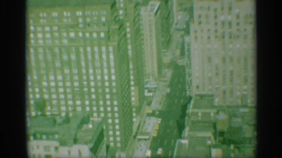 1959 New York home-movie frame showing high-rise buildings, gridded windows, and a narrow Manhattan street from above.