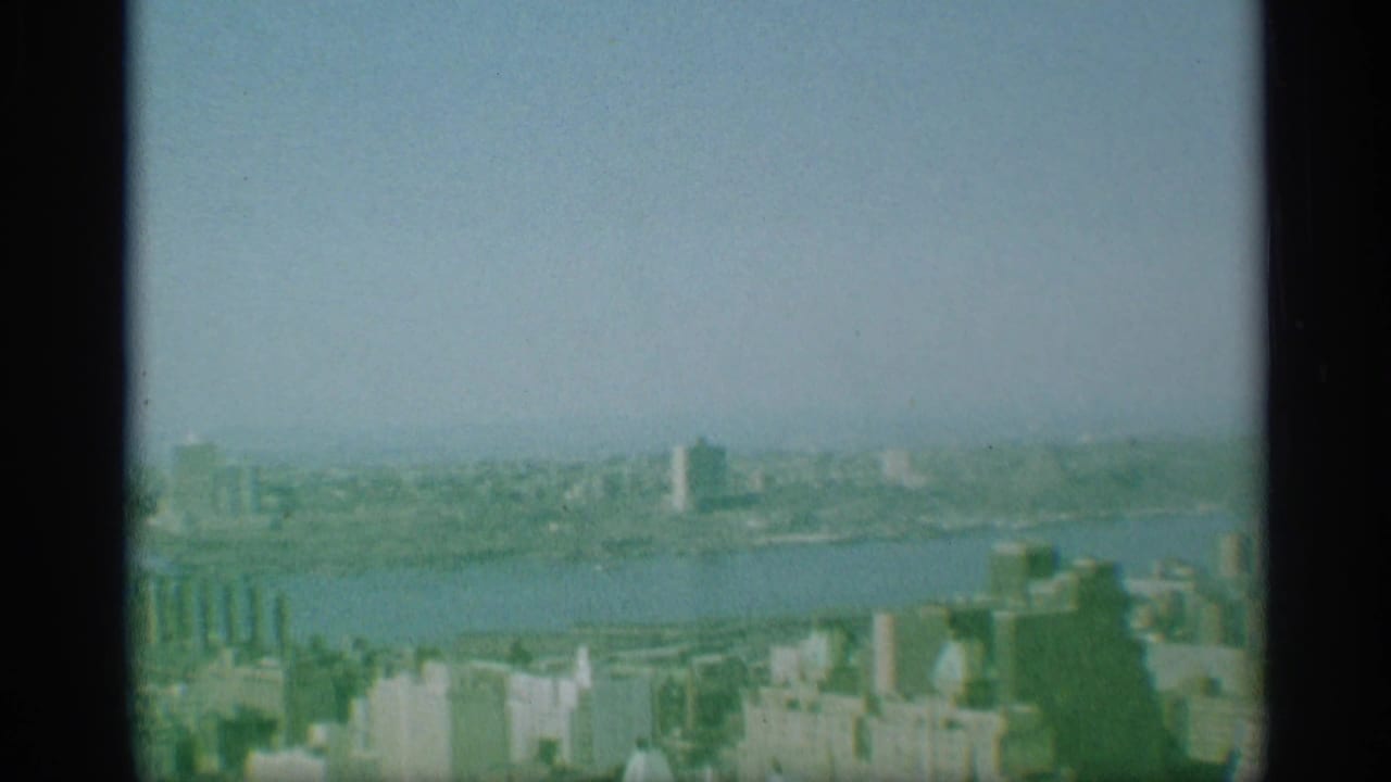 1959 New York 8mm frame showing a hazy aerial view over a river, waterfront structures, and distant city buildings.