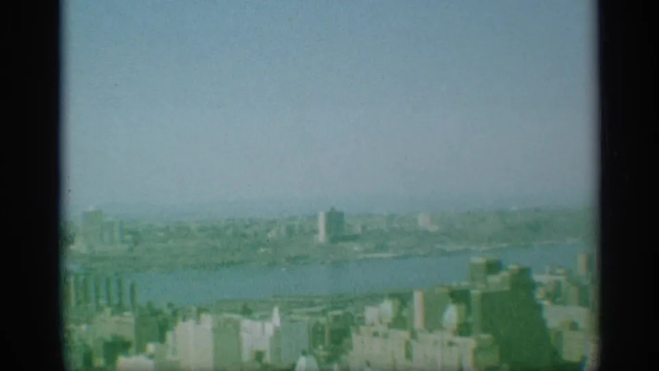1959 New York 8mm frame showing a hazy aerial view over a river, waterfront structures, and distant city buildings.