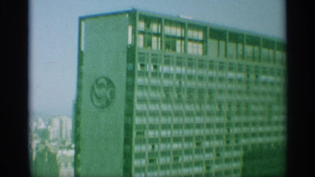1959 New York aerial frame shows a tall high-rise with a visible circular logo, with distant buildings and sky behind it.