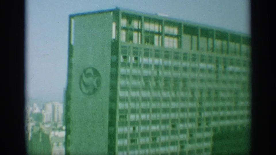 1959 New York aerial frame shows a tall high-rise with a visible circular logo, with distant buildings and sky behind it.