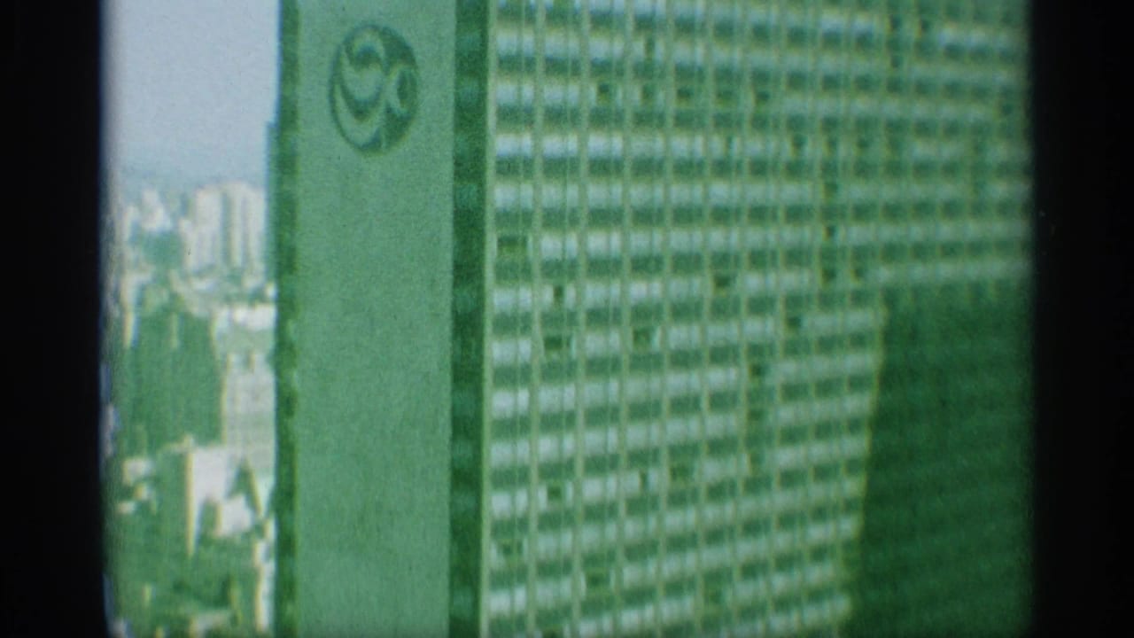 1959 New York 8mm frame showing a tall mid-century building facade with repeated windows, pale vertical bands, dark gate edges, and a small logo-like mark.