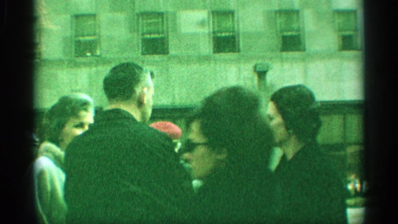 1959 New York home-movie frame with bundled pedestrians moving past a large building facade on a busy sidewalk.