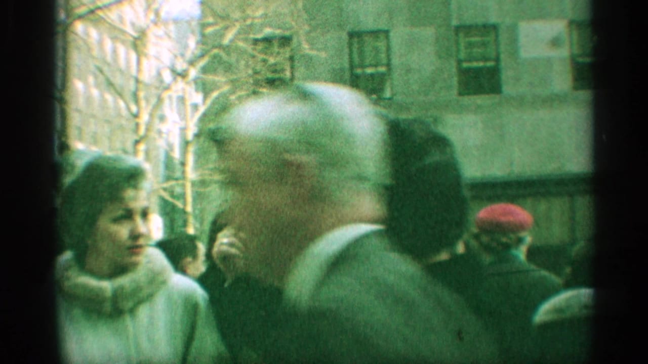1959 New York street frame with a balding man crossing the foreground and other pedestrians near a building facade.