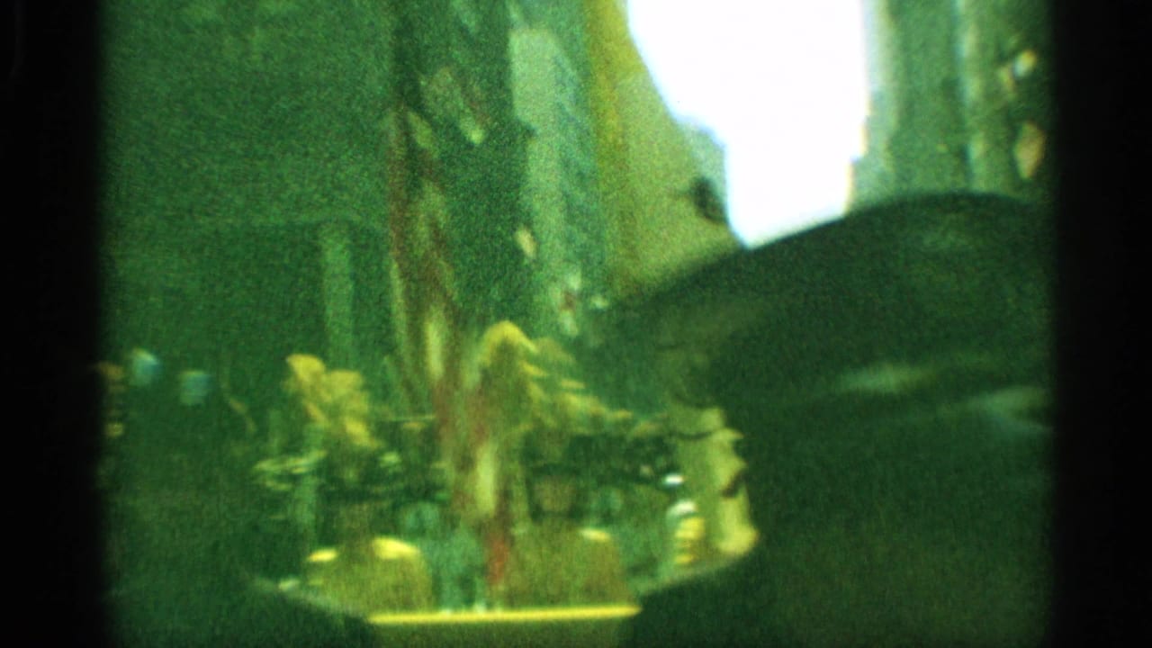 1959 New York parade frame with a drum major or uniformed leader near flags, crowds, and tall buildings in soft handheld 8mm focus.