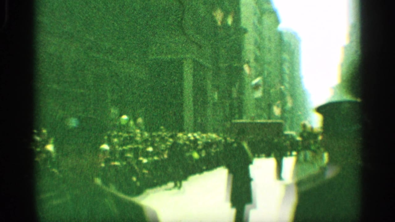 1959 New York parade frame showing marching band musicians moving past sidewalk spectators, with tall buildings and heavy green 8mm grain.