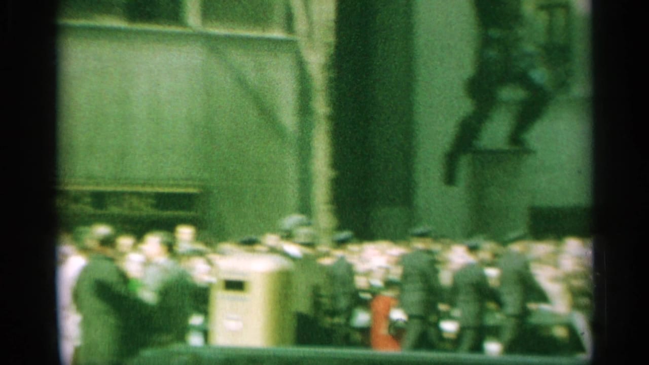 Grainy 1959 New York street frame with spectators, officers, parked cars, and a wall-mounted figure detail.