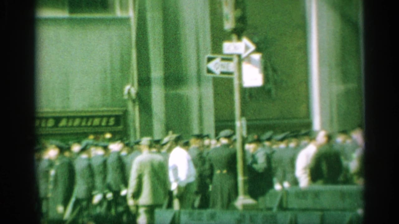 In 1959 New York, uniformed men in dark suits and hats pass a building with a Trans World Airlines sign as pedestrians crowd the foreground.