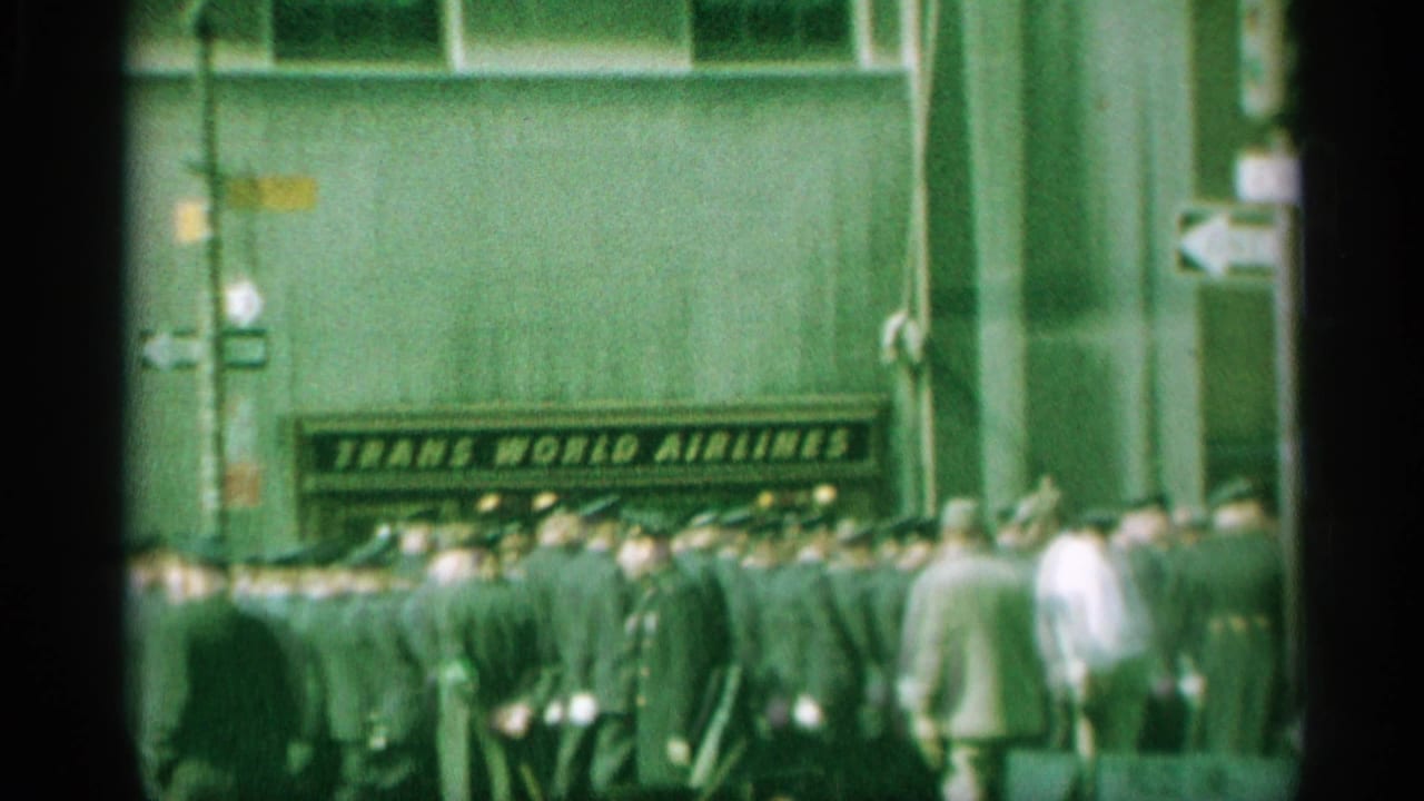 A crowded 1959 New York street view shows uniformed men in formation moving past a visible Trans World Airlines sign and street-direction markers.