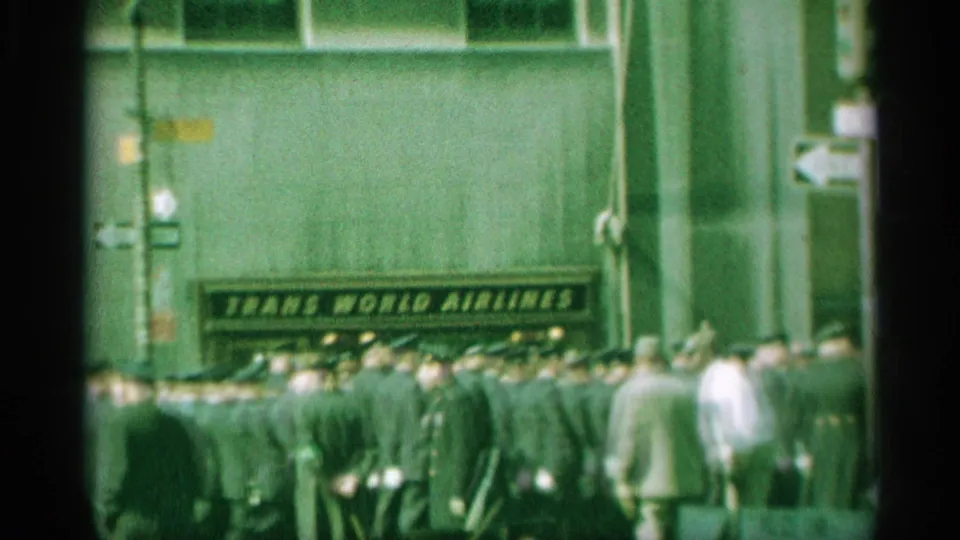 A crowded 1959 New York street view shows uniformed men in formation moving past a visible Trans World Airlines sign and street-direction markers.