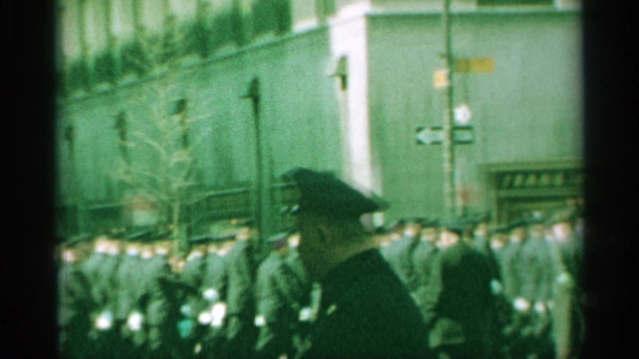 The camera catches a blurred 1959 New York street crowd, with uniformed men in formation, a nearby officer-like silhouette, and the TWA sign partly visible.