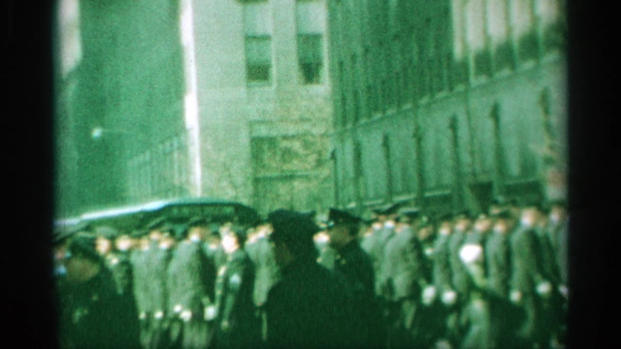 Uniformed men march in formation along a city street between tall buildings.