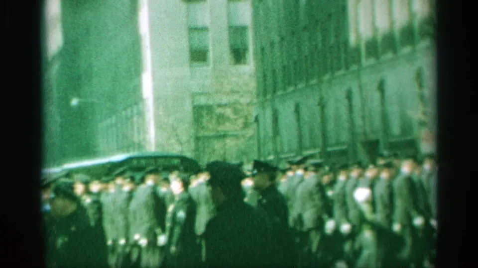 1959 New York home-movie frame showing uniformed men marching in formation on a city street, partly soft and green-tinted.