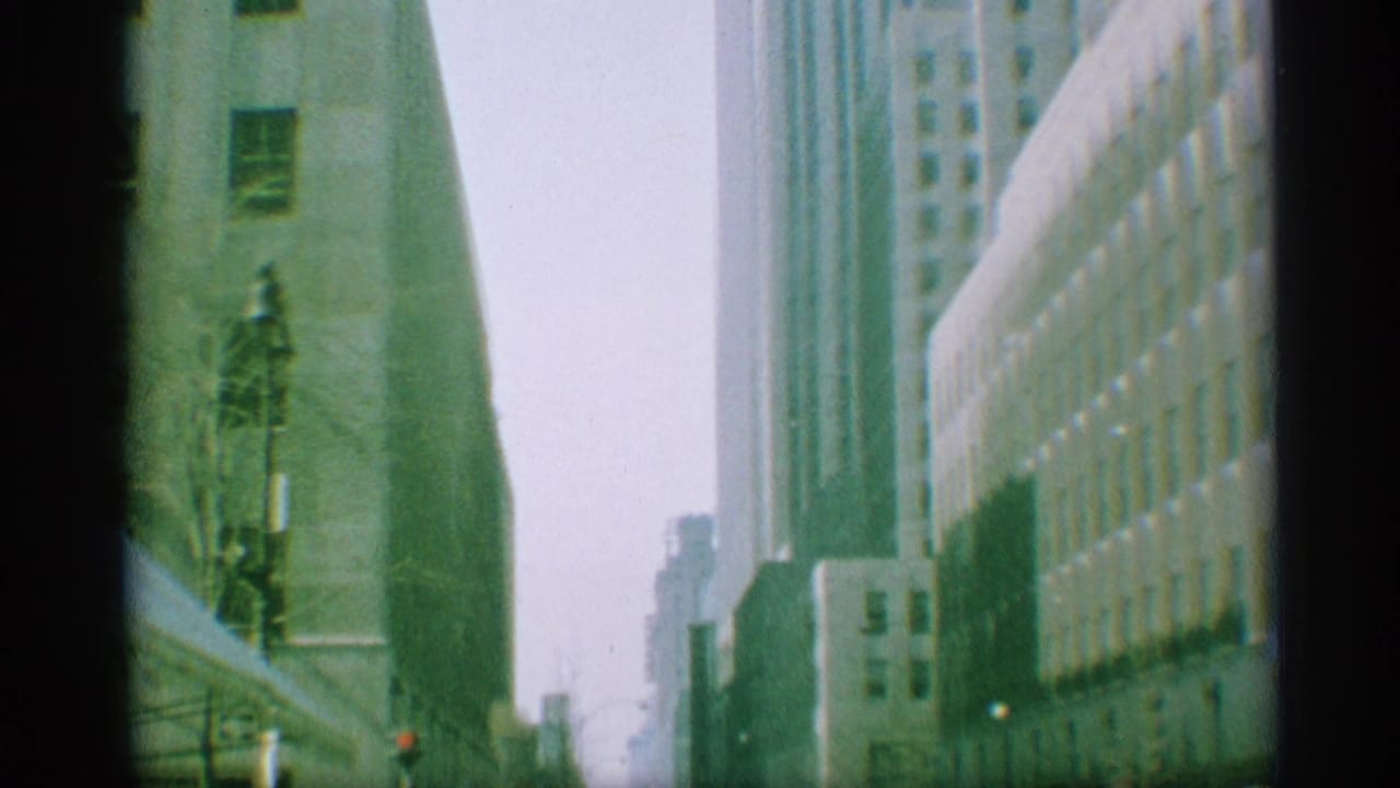 1959 New York frame looking down a built-up city street with high-rises, a streetlamp, and soft green 8mm color.