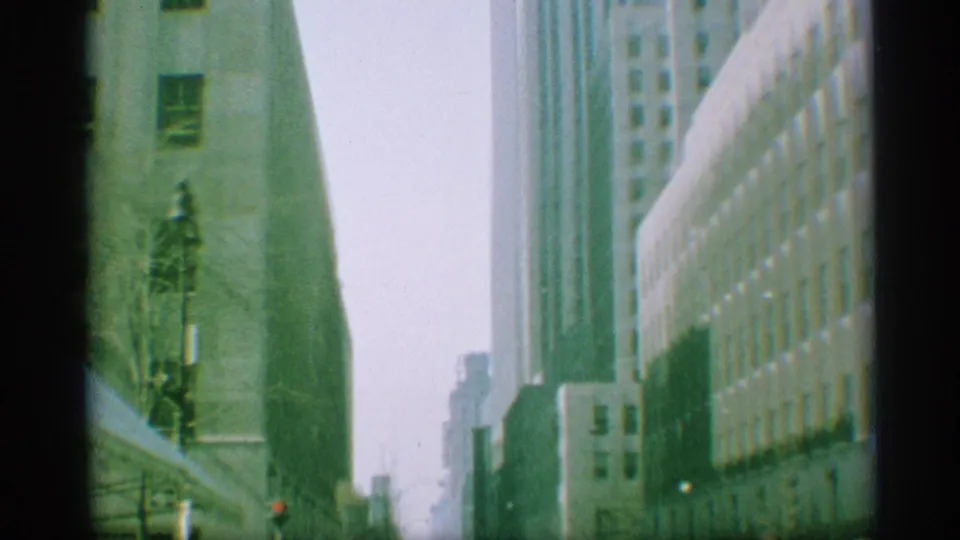 1959 New York frame looking down a built-up city street with high-rises, a streetlamp, and soft green 8mm color.