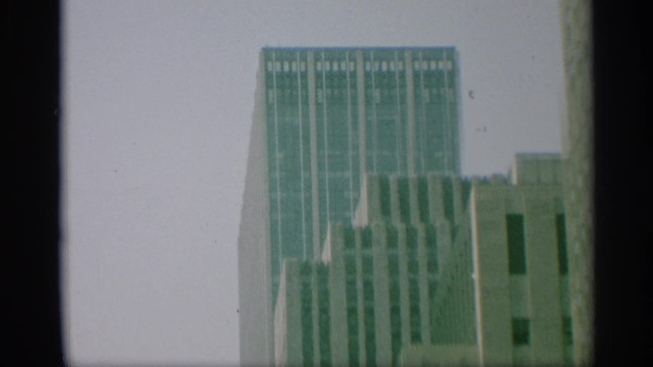1959 New York 8mm frame showing a tall vertical skyscraper beside neighboring upper-story facades, all softened by film grain.