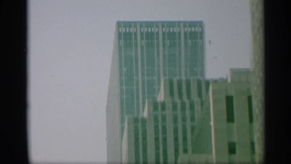 1959 New York 8mm frame showing a tall vertical skyscraper beside neighboring upper-story facades, all softened by film grain.