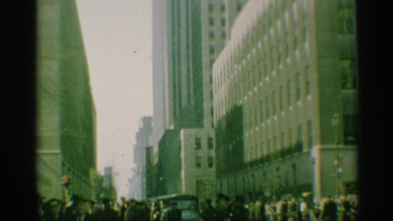1959 New York street scene with pedestrians and vehicles beneath tall buildings, filmed from a low viewpoint.