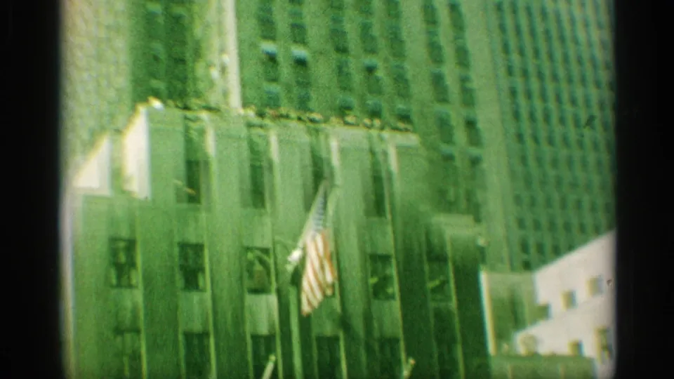 1959 New York frame showing an American flag on a building facade amid neighboring high-rises.