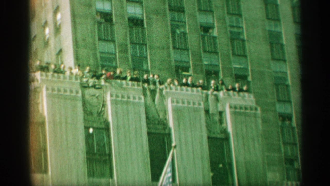 1959 New York frame with distant people gathered along a rooftop ledge above a tall urban facade and American flag.