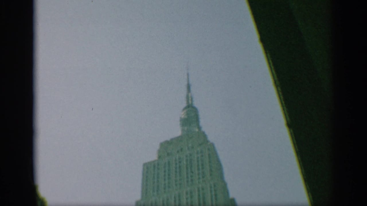 1959 New York home-movie frame with the Empire State Building crown smaller in the frame beneath a pale sky and dark edge shadow.