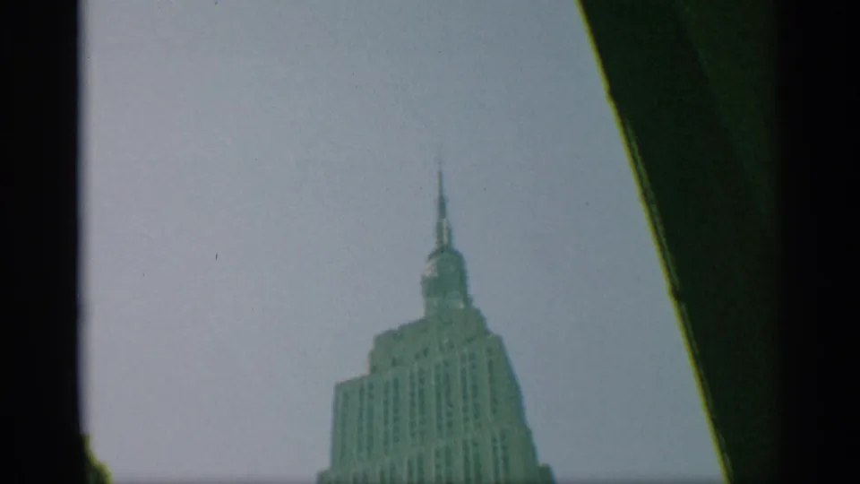 1959 New York home-movie frame with the Empire State Building crown smaller in the frame beneath a pale sky and dark edge shadow.