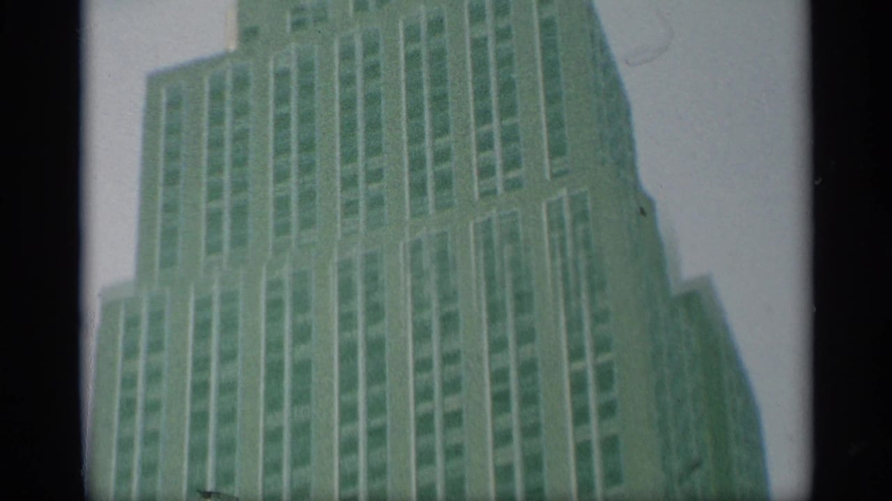 1959 New York home-movie frame looking up at a tall setback skyscraper with rows of windows and soft green film color shift.