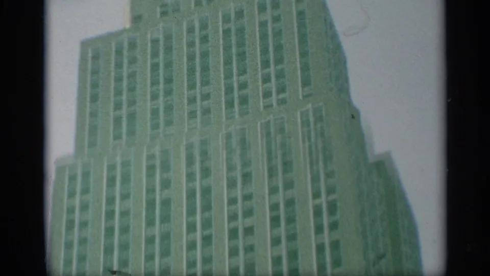 1959 New York home-movie frame looking up at a tall setback skyscraper with rows of windows and soft green film color shift.