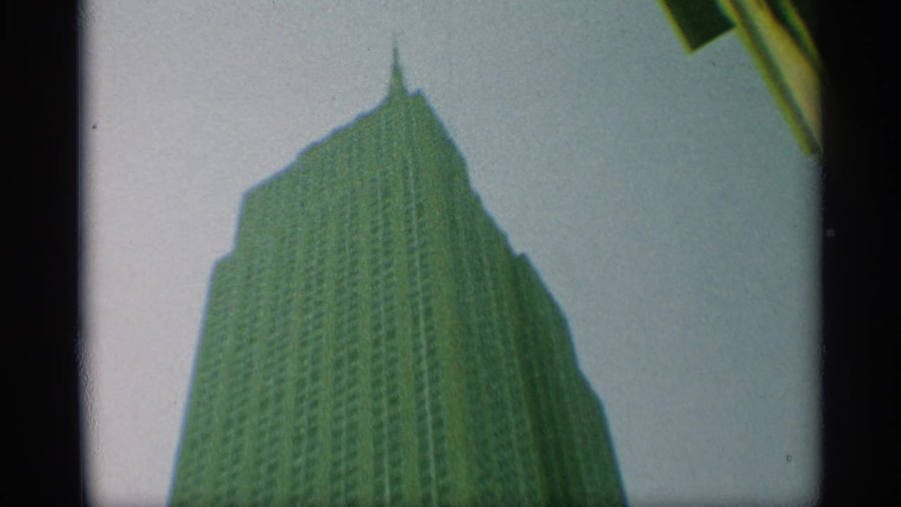 Street-level home-movie frame of a tall New York skyscraper rising to a pointed upper section against open sky.