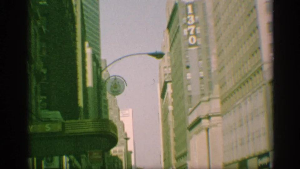 1959 New York 8mm view looking up a city street toward high-rise facades, a vertical 1370 sign, and a curved street lamp.