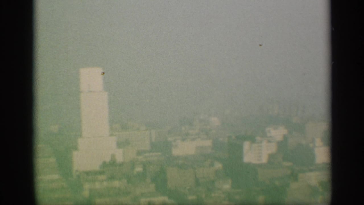 Color-faded 8mm skyline view in New York, showing a pale tower form and distant buildings through haze or smog.