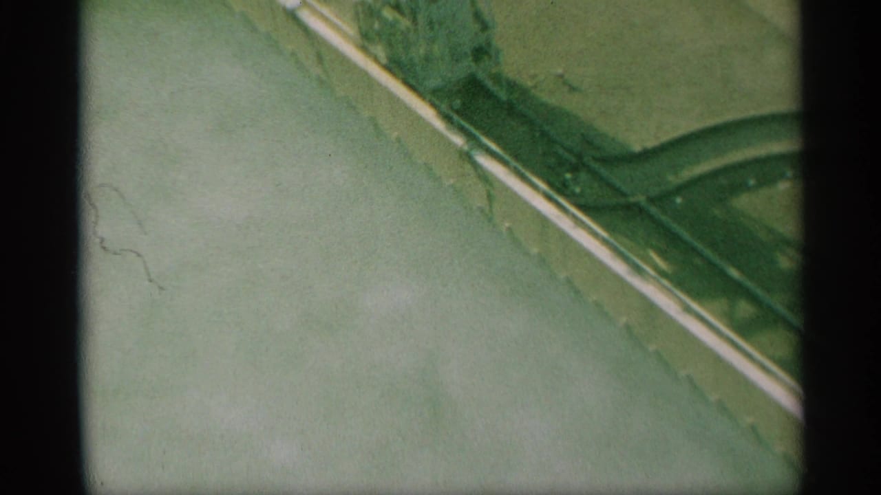 1959 New York 8mm frame looking down at a paved street corner, white curb line, and green metal fencing from an elevated position.