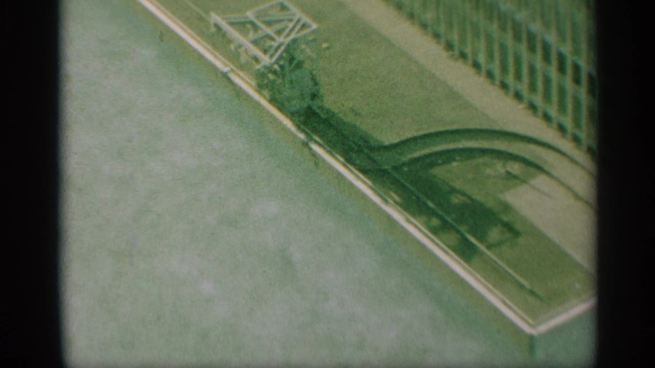 1959 New York 8mm frame showing a high-angle street corner with green railing, pale curb edging, and a grid-like structure at the side.