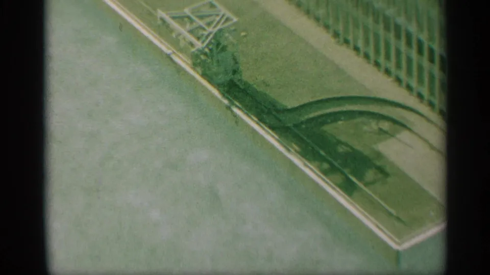 1959 New York 8mm frame showing a high-angle street corner with green railing, pale curb edging, and a grid-like structure at the side.