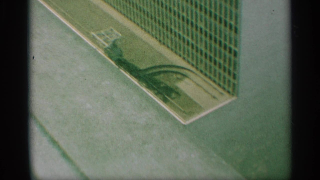 1959 New York 8mm frame looking down on a paved corner, green fence panels, white curb markings, and a small light-toned object near the curb.