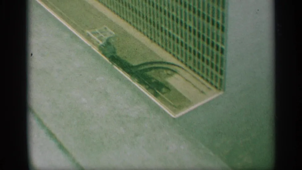 1959 New York 8mm frame looking down on a paved corner, green fence panels, white curb markings, and a small light-toned object near the curb.