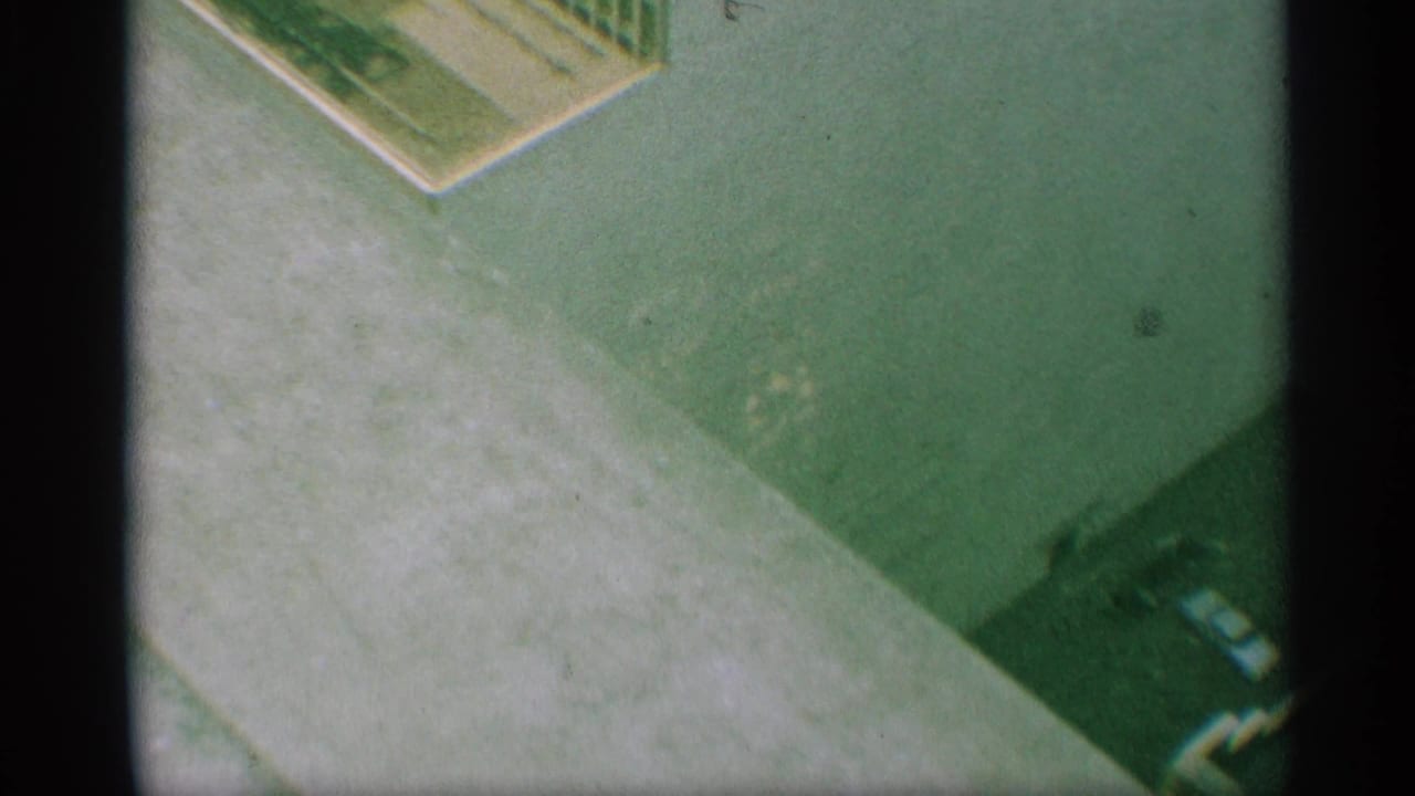1959 New York 8mm frame showing a high-angle paved area near a building edge, with curb line, deep shadow, and a partial dark vehicle-like shape.