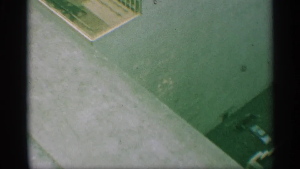 1959 New York 8mm frame showing a high-angle paved area near a building edge, with curb line, deep shadow, and a partial dark vehicle-like shape.
