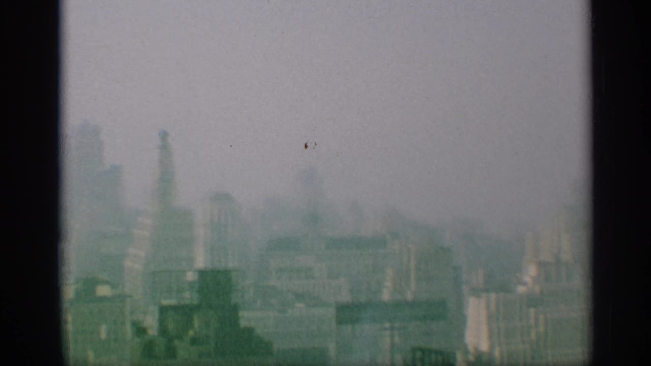 A hazy 1959 New York urban view shows tall towers and lower buildings softened by 8mm grain and exposure.