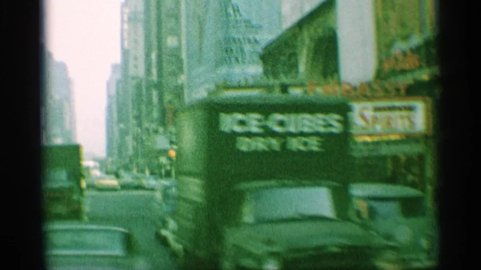 1959 New York street view with automobiles, pedestrians, storefront signage, and a theater marquee seen from a handheld 8mm camera.