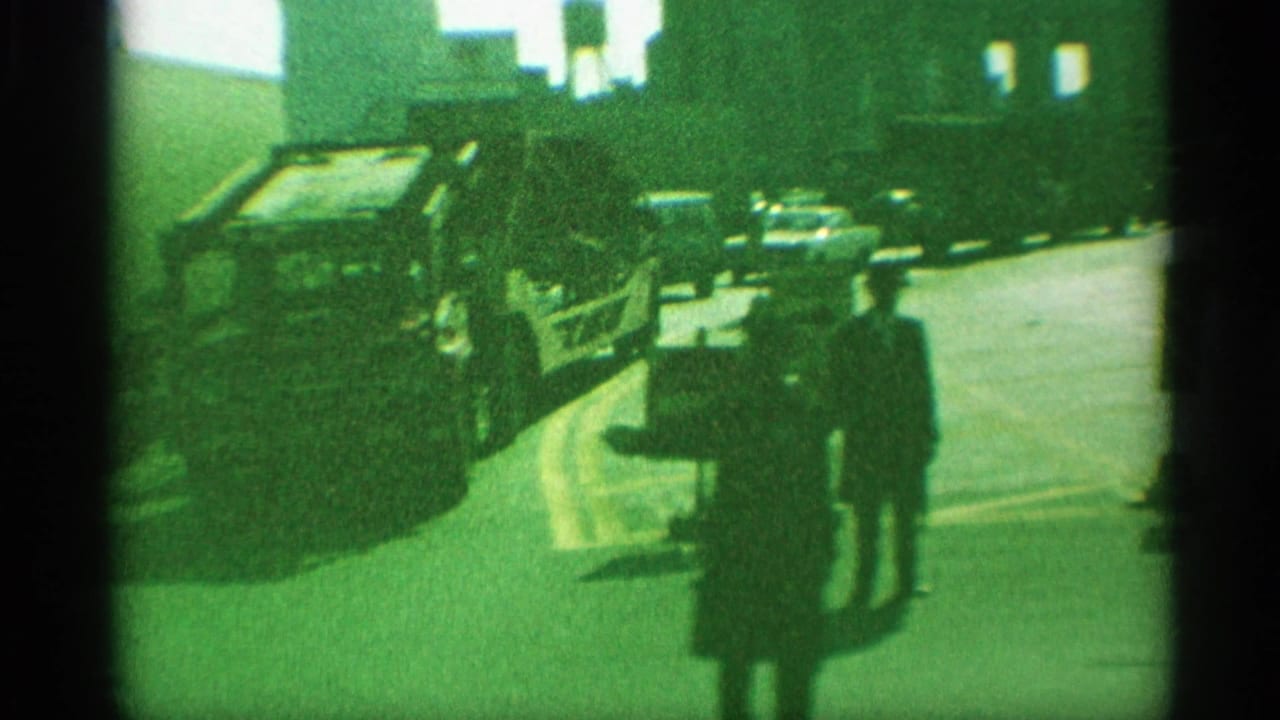 1959 New York 8mm frame with pedestrians on the sidewalk, vintage cars along the curb, and city buildings in shadow.