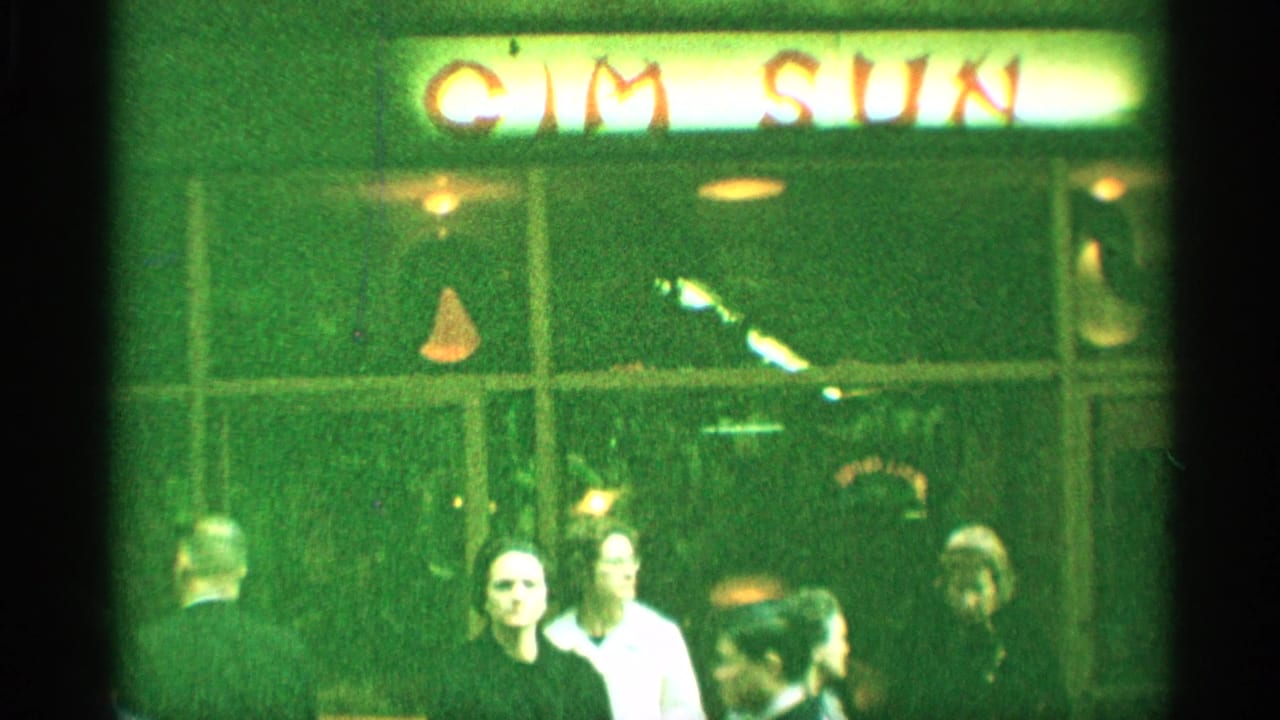1959 New York 8mm frame showing adults gathered outside a green-lit storefront with a Cim Sun sign overhead.