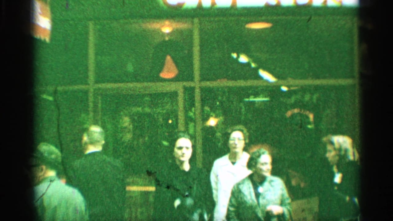 1959 New York home-movie frame showing adults outside a green-lit storefront, with the sign partly cropped above.