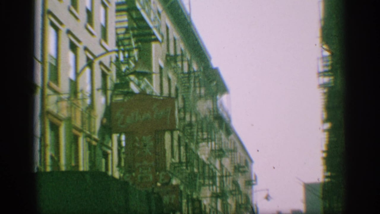 1959 New York street view with walk-up buildings, fire escapes, and a sunlit roadway receding into the distance.