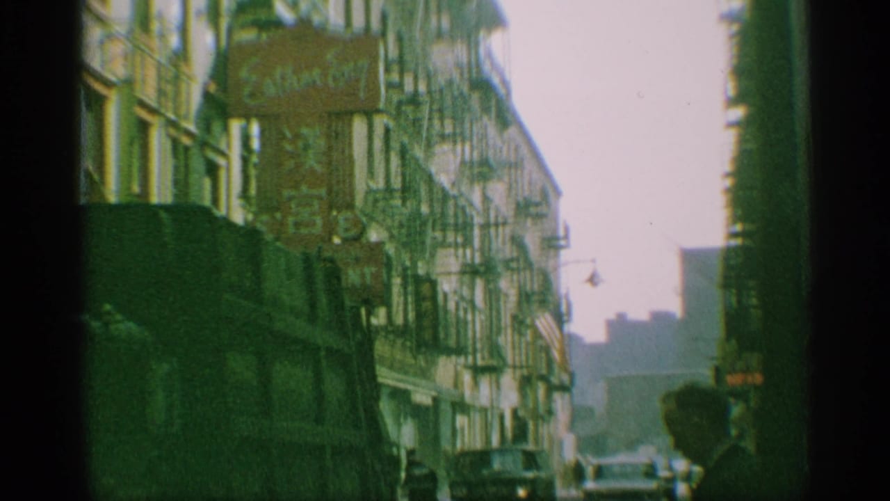 1959 New York 8mm street frame showing fire escapes, parked or moving cars, and a shadowed passerby or foreground figure.