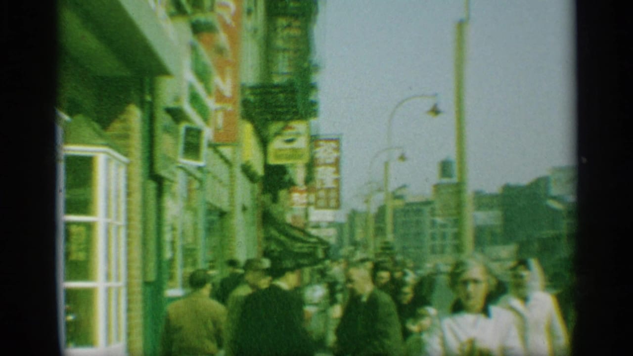 1959 New York sidewalk view with pedestrians moving past storefronts, hanging signs, fire escapes, and mid-century street fixtures.